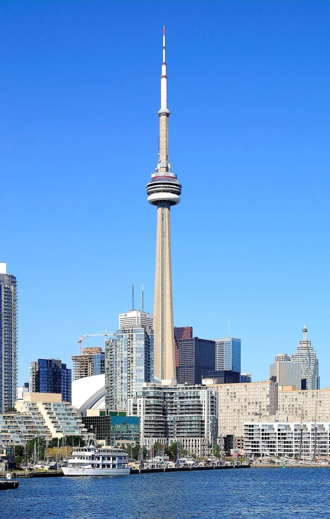 Vista della città di Toronto in Canada. Il cielo è sereno e al centro svetta imperiosa la CN Tower, tutta bianco panna. Tutto attorno si sviluppano altri palazzi e vie cittadine. Ai piedi della torre si può vedere parte del lago Ontario
